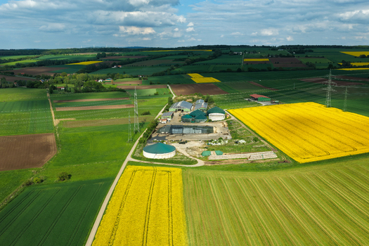 Aerial view of a biogas plant and farm buildings surrounded by vibrant yellow rapeseed fields and green agricultural land under a cloudy sky in Hofgeismar Hessen Germany. Aerial view of a biogas plant and farm buildings surrounded by vibrant yellow rapeseed fields and green agricultural land under a cloudy sky in Hofgeismar Hessen Germany.
