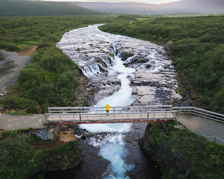 Aerial view of Bruarfoss waterfall with blue glacial water flowing through dark rocks and a person in a yellow jacket on a wooden bridge in Blaskogabyggd Iceland. Aerial view of Bruarfoss waterfall with blue glacial water flowing through dark rocks and a person in a yellow jacket on a wooden bridge in Blaskogabyggd Iceland.