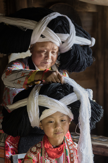 Gaoxing China 29 April 2026 View of an older woman dressing a young girl in traditional Long Horn Miao attire with a large black and white yarn headpiece in a wooden interior. Gaoxing China 29 April 2026 View of an older woman dressing a young girl in traditional Long Horn Miao attire with a large black and white yarn headpiece in a wooden interior.