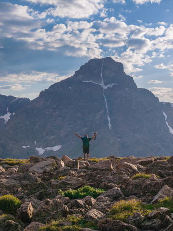 Aerial view of a hiker standing on a rocky ridge with arms raised towards the Mount of the Holy Cross surrounded by alpine terrain in Vail Colorado United States. Aerial view of a hiker standing on a rocky ridge with arms raised towards the Mount of the Holy Cross surrounded by alpine terrain in Vail Colorado United States.