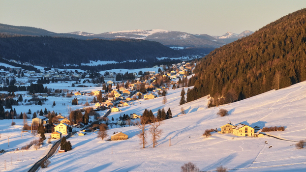 Aerial view of a snow covered village nestled between dark forests and rolling hills bathed in the warm glow of the setting sun Les Rousses Bourgogne Franche Comté France. Aerial view of a snow covered village nestled between dark forests and rolling hills bathed in the warm glow of the setting sun Les Rousses Bourgogne Franche Comté France.
