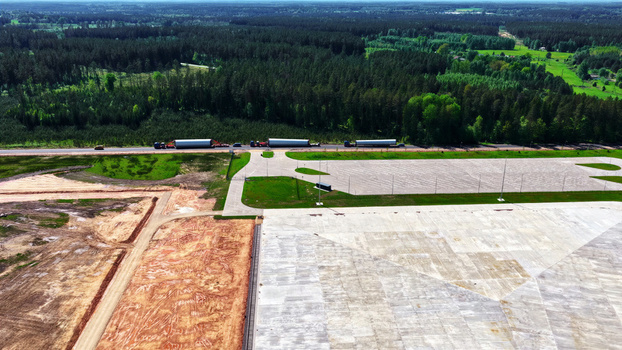 Aerial view of trucks transporting wind turbine blades along a road next to a large concrete construction site and dense forest in Latvia. Aerial view of trucks transporting wind turbine blades along a road next to a large concrete construction site and dense forest in Latvia.