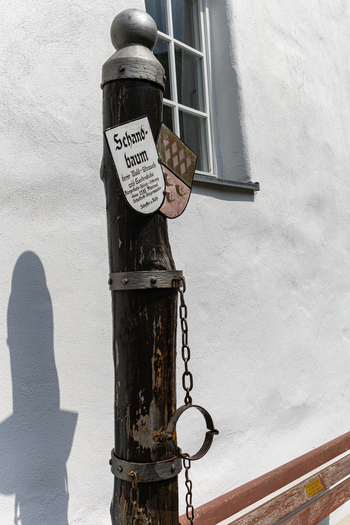 View of the historic Schandbaum pillory with iron shackles and a coat of arms against a white building wall in Monreal Rhineland Palatinate Germany. View of the historic Schandbaum pillory with iron shackles and a coat of arms against a white building wall in Monreal Rhineland Palatinate Germany.