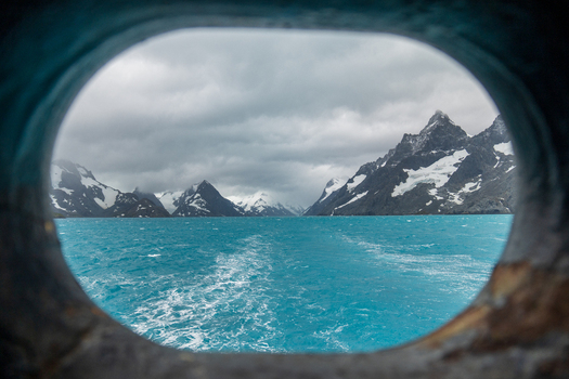 View of Drygalski Fjord with turquoise glacial water and snow capped mountains under a cloudy sky seen through a ship porthole South Georgia and the South Sandwich Islands.