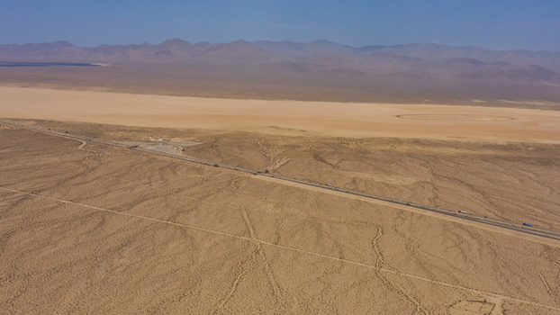 Aerial view of a vast desert landscape with a long highway stretching across the arid terrain featuring a dry lake bed and distant mountains under a clear blue sky in the United States.