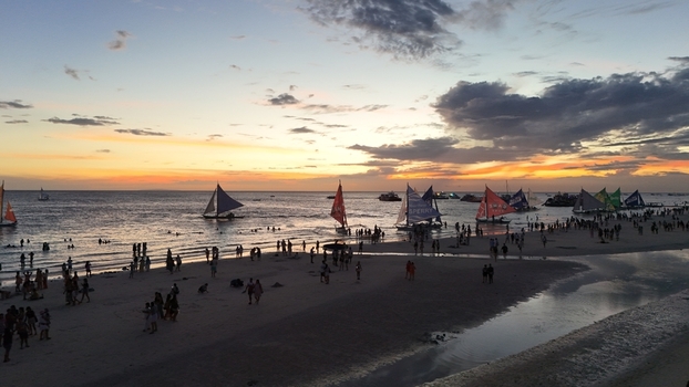 Malay Philippines 02 May 2026 Aerial view of Boracay Island beach at sunset with numerous traditional paraw sailboats and people gathered on the white sand under a dramatic orange sky.