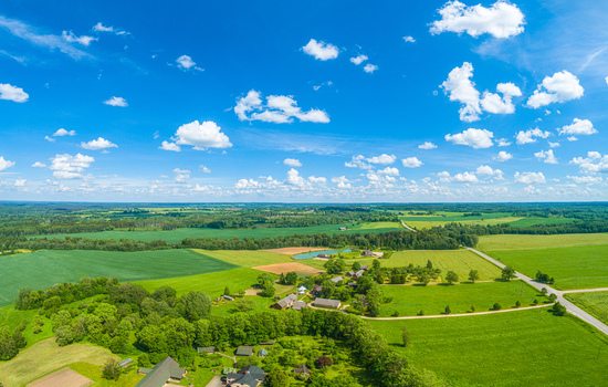 Aerial view of lush green fields agricultural land small rural houses and a small pond under a bright blue sky with scattered white clouds in Latvia. Aerial view of lush green fields agricultural land small rural houses and a small pond under a bright blue sky with scattered white clouds in Latvia.