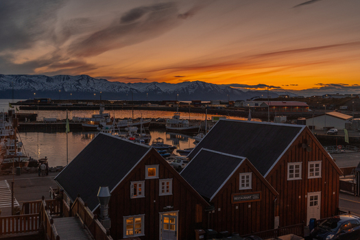 View of the harbor reflecting the fiery sunset sky contrasted against the dark wooden buildings and distant snow capped mountains Húsavík Norðurþing Iceland. View of the harbor reflecting the fiery sunset sky contrasted against the dark wooden buildings and distant snow capped mountains Húsavík Norðurþing Iceland.