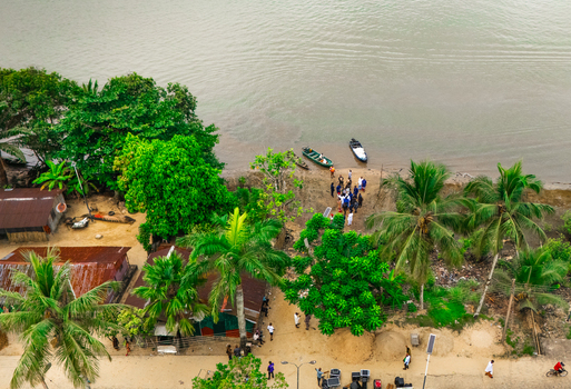 Aerial view of people gathered near small boats on the riverbank surrounded by lush palm trees and simple structures in Minima Rivers Nigeria.