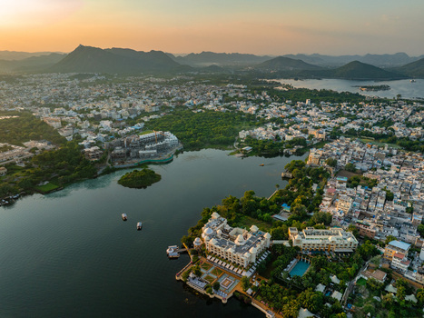 Aerial view of Lake Pichola and the historic city featuring grand palaces lush gardens and calm waters under a warm sunset sky Udaipur Rajasthan India.