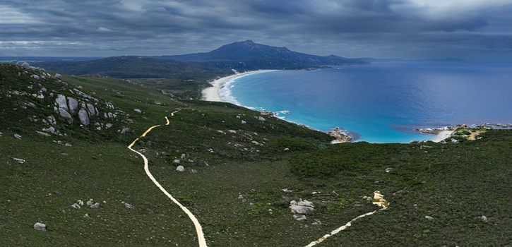 Aerial view of Bettys Beach featuring a winding dirt path through lush green coastal vegetation turquoise waters and rocky outcrops under a dramatic cloudy sky in Albany WA Australia. Aerial view of Bettys Beach featuring a winding dirt path through lush green coastal vegetation turquoise waters and rocky outcrops under a dramatic cloudy sky in Albany WA Australia.
