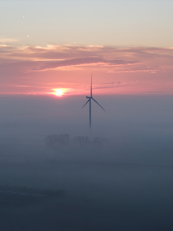 Aerial view of a single wind turbine rising above a thick layer of morning fog under a vibrant pink and orange sunrise sky in Vrouwenpolder Zeeland Netherlands.