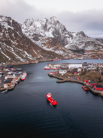 Aerial view of a deep blue fjord embraced by rugged snow dusted mountains and dotted with red buildings the scene punctuated by a bright red ship Ballstad Nordland Norway. Aerial view of a deep blue fjord embraced by rugged snow dusted mountains and dotted with red buildings the scene punctuated by a bright red ship Ballstad Nordland Norway.