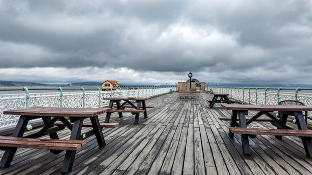 View of Mumbles Pier featuring wooden picnic tables and ornate white railings under a dramatic overcast sky in Mumbles Wales United Kingdom.