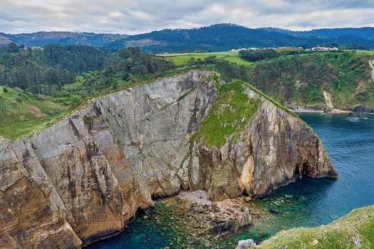Aerial view of Silence Beach Viewpoint featuring dramatic jagged cliffs turquoise ocean water and lush green hills under a cloudy sky in Cudillero Asturias Spain.