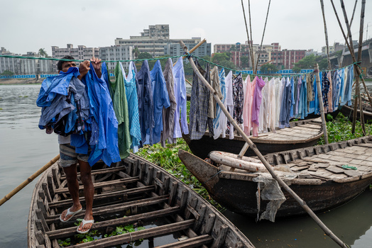 Dhaka Division Bangladesh 14 May 2026 View of a man hanging laundry on a line across traditional wooden boats on the Buriganga River with the city skyline in the background.