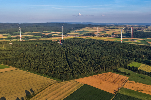 Aerial view of a dense forest surrounded by agricultural fields and wind turbines under a clear blue sky in Hofgeismar Hessen Germany.