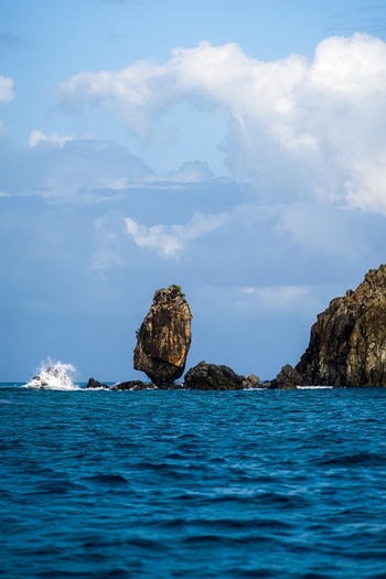 View of a tall sea stack rising from the blue ocean with waves crashing against rocks under a cloudy sky in Fernando de Noronha State of Pernambuco Brazil. View of a tall sea stack rising from the blue ocean with waves crashing against rocks under a cloudy sky in Fernando de Noronha State of Pernambuco Brazil.