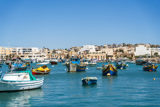 View of traditional colorful luzzu fishing boats moored in the calm blue waters of the harbor with the coastal town in the background in Marsaskala Southern Region Malta. View of traditional colorful luzzu fishing boats moored in the calm blue waters of the harbor with the coastal town in the background in Marsaskala Southern Region Malta.