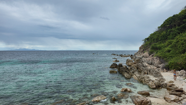 View of the rocky coastline and turquoise waters of Ko Tao with a lush green hillside under a cloudy sky in Ko Tao Surat Thani Thailand.