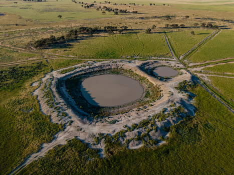 Aerial view of a large circular muddy water hole surrounded by green agricultural fields and sparse trees in the flat landscape of Hay New South Wales Australia.
