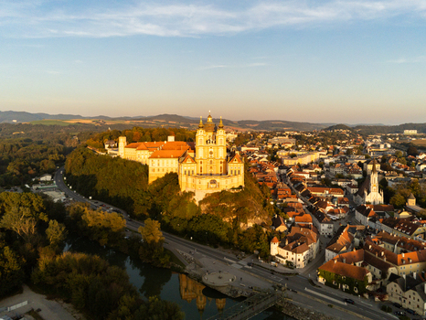 Aerial view of Melk Abbey illuminated by golden hour light featuring historic architecture surrounding town buildings and lush greenery along the river in Melk Niederosterreich Austria. Aerial view of Melk Abbey illuminated by golden hour light featuring historic architecture surrounding town buildings and lush greenery along the river in Melk Niederosterreich Austria.