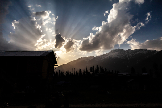 View of bright sun rays breaking through clouds over snow capped mountains and silhouetted buildings in the Neelum Valley Arang Kel Azad Kashmir Pakistan. View of bright sun rays breaking through clouds over snow capped mountains and silhouetted buildings in the Neelum Valley Arang Kel Azad Kashmir Pakistan.