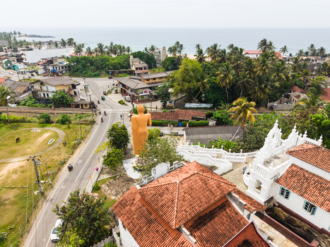 Aerial view of the Kumarakanda Kumara Maha Viharaya temple featuring a large golden Buddha statue white buildings and lush palm trees in Hikkaduwa Southern Province Sri Lanka. Aerial view of the Kumarakanda Kumara Maha Viharaya temple featuring a large golden Buddha statue white buildings and lush palm trees in Hikkaduwa Southern Province Sri Lanka.