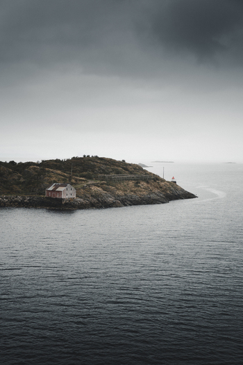 View of a small red cabin on a rocky coastal cliff with a lighthouse and wooden drying racks under a moody overcast sky in Henningsvaer Nordland Norway. View of a small red cabin on a rocky coastal cliff with a lighthouse and wooden drying racks under a moody overcast sky in Henningsvaer Nordland Norway.