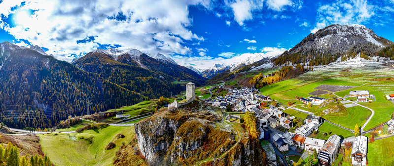 Aerial view of the historic village of Ardez and Steinsberg Castle ruins perched on a rocky hill surrounded by autumn forests and snow capped peaks in Scuol Grisons Switzerland.