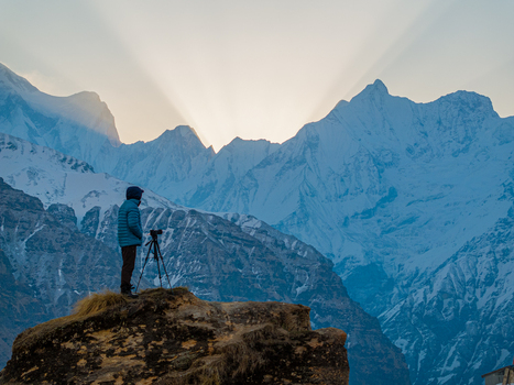 Aerial view of a photographer with a camera on a tripod standing on a rocky outcrop overlooking the snow capped Himalayan mountains at sunrise in Nepal.
