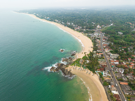 Aerial view of the sandy coastline with turquoise water rocky outcrops and a coastal road lined with tropical trees in Dodanduwa Southern Province Sri Lanka. Aerial view of the sandy coastline with turquoise water rocky outcrops and a coastal road lined with tropical trees in Dodanduwa Southern Province Sri Lanka.