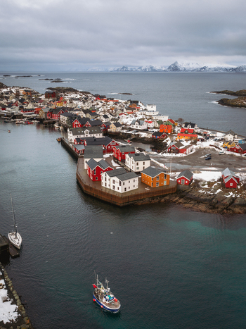 Aerial view of vibrant red and white buildings nestled along the rugged coastline contrasting with the dark waters and snow dusted peaks Henningsv&aelig;r Nordland Norway.