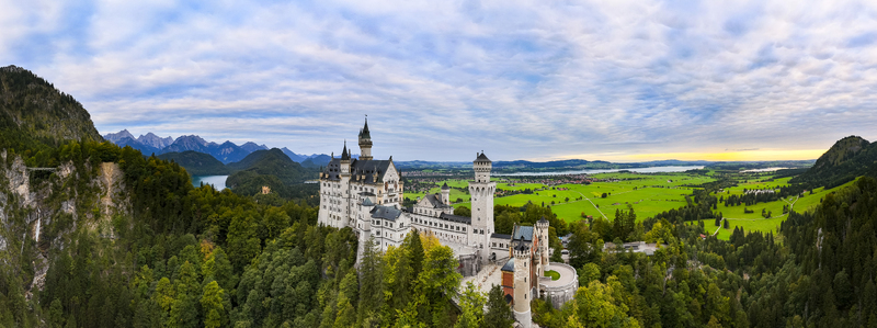 Aerial view of Neuschwanstein Castle s pale stone and spires rise amidst lush forests juxtaposed against the distant Alpine peaks Füssen Bayern Germany. Aerial view of Neuschwanstein Castle s pale stone and spires rise amidst lush forests juxtaposed against the distant Alpine peaks Füssen Bayern Germany.