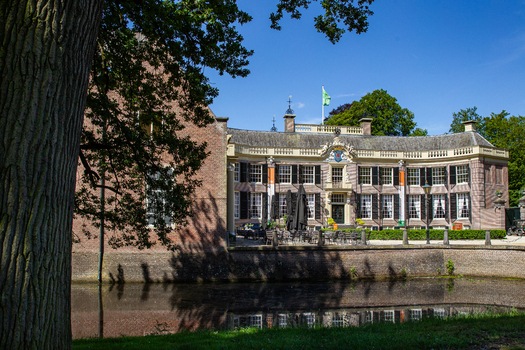 View of Castle Groeneveld with its historic brick facade and moat surrounded by lush green trees under a clear blue sky in Baarn Utrecht Netherlands.