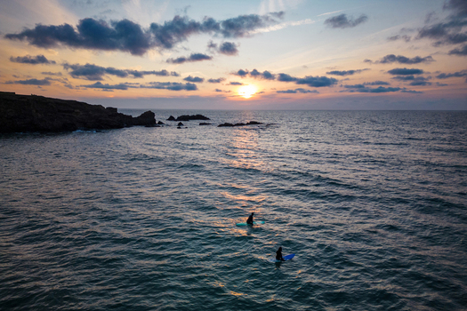 Aerial view of two surfers on the water near the rocky coastline at sunset with a cloudy sky Bude England United Kingdom.