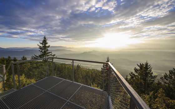 View of sunlight piercing through the clouds over the mountains seen from a metal platform surrounded by lush trees Brandlalm Styria Austria. View of sunlight piercing through the clouds over the mountains seen from a metal platform surrounded by lush trees Brandlalm Styria Austria.