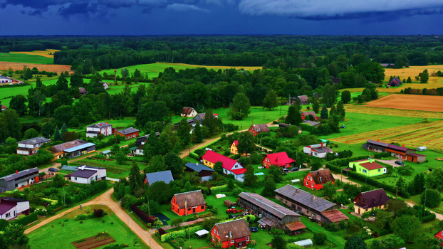 Aerial view of a rural village featuring colorful houses with red roofs lush green fields and dirt roads under a dramatic dark stormy sky in Latvia. Aerial view of a rural village featuring colorful houses with red roofs lush green fields and dirt roads under a dramatic dark stormy sky in Latvia.