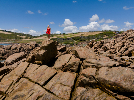 Aerial view of a woman in a red dress standing on the rocky coastline at Injidup Natural Spa with green hills and blue sky in Yallingup Western Australia Australia. Aerial view of a woman in a red dress standing on the rocky coastline at Injidup Natural Spa with green hills and blue sky in Yallingup Western Australia Australia.
