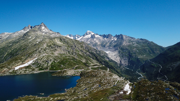 Aerial view of the Grimsel Pass with winding mountain roads a deep blue lake and rugged rocky peaks under a clear sky in Obergoms Valais Switzerland. Aerial view of the Grimsel Pass with winding mountain roads a deep blue lake and rugged rocky peaks under a clear sky in Obergoms Valais Switzerland.