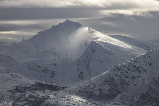 View of majestic snow covered mountain peaks under a dramatic cloudy sky with sunlight illuminating the summit Tromso Troms Norway.