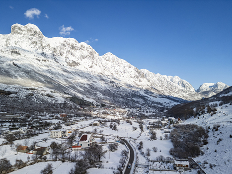Aerial view of a snow covered mountain village nestled in the Albanian Alps with winding roads scattered houses and dramatic peaks under a clear blue sky in Boge Shkoder County Albania. Aerial view of a snow covered mountain village nestled in the Albanian Alps with winding roads scattered houses and dramatic peaks under a clear blue sky in Boge Shkoder County Albania.