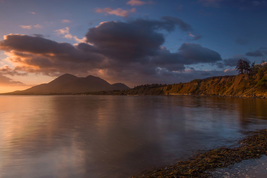 View of Lettergesh Beach with calm water reflecting dramatic sunset clouds and distant mountains in County Galway Ireland.