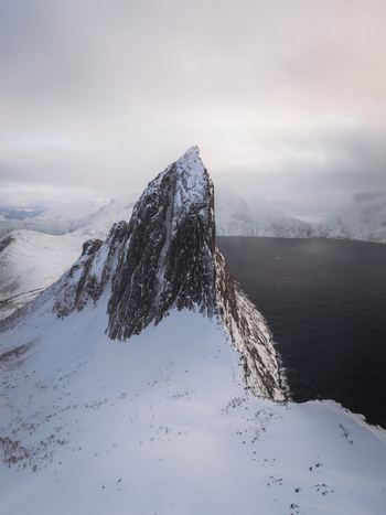 Aerial view of the stark snow laden peak of the Okshornan mountains contrasting against the still dark waters of the fjord Fjordgård Troms Norway. Aerial view of the stark snow laden peak of the Okshornan mountains contrasting against the still dark waters of the fjord Fjordgård Troms Norway.