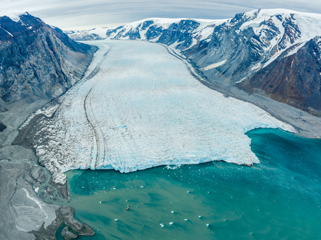 Aerial view of a massive glacier flowing into a turquoise glacial lake surrounded by rugged snow capped mountains and rocky terrain in Greenland.