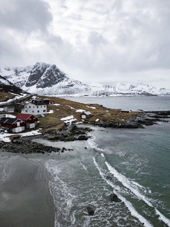 Aerial view of the coastal village of Tromvik near Grotfjord with traditional red houses snow capped mountains and waves crashing on a sandy beach Tromvik Troms Norway. Aerial view of the coastal village of Tromvik near Grotfjord with traditional red houses snow capped mountains and waves crashing on a sandy beach Tromvik Troms Norway.