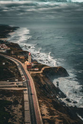 Aerial view of Farol Penedo da Saudade lighthouse on a rugged cliff overlooking the Atlantic Ocean with crashing waves and a coastal road in Marinha Grande Leiria Portugal. Aerial view of Farol Penedo da Saudade lighthouse on a rugged cliff overlooking the Atlantic Ocean with crashing waves and a coastal road in Marinha Grande Leiria Portugal.