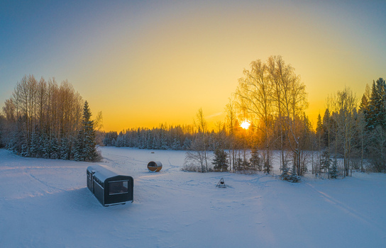 Aerial view of a small cabin and a barrel sauna in a snow covered field surrounded by a forest during a golden sunset in Latvia. Aerial view of a small cabin and a barrel sauna in a snow covered field surrounded by a forest during a golden sunset in Latvia.