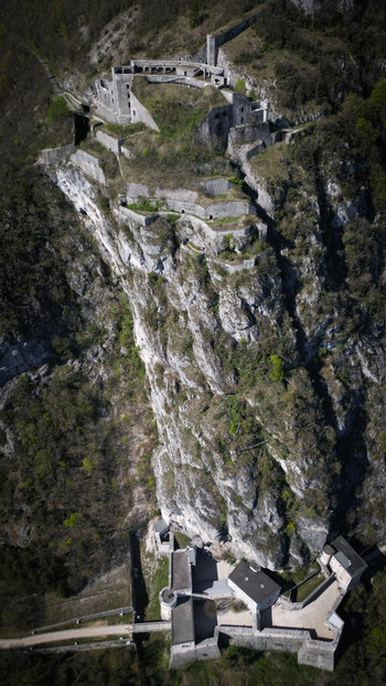 Aerial view of the stark white ruins of Fort l &Eacute;cluse clinging to the rugged green clad cliffs a testament to history s enduring presence L&eacute;az Auvergne Rh&ocirc;ne Alpes France.
