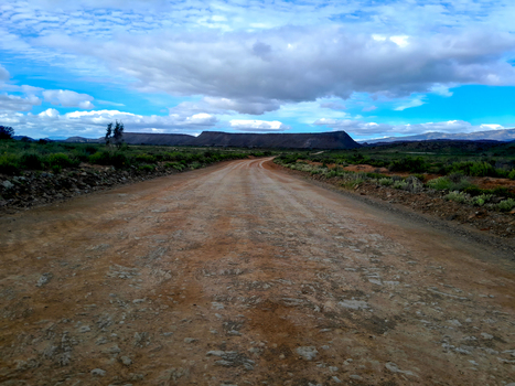 View of a gravel road stretching through the semi arid plains of the Anysberg Nature Reserve with rugged mountains under a blue sky in Anysberg Western Cape South Africa. View of a gravel road stretching through the semi arid plains of the Anysberg Nature Reserve with rugged mountains under a blue sky in Anysberg Western Cape South Africa.
